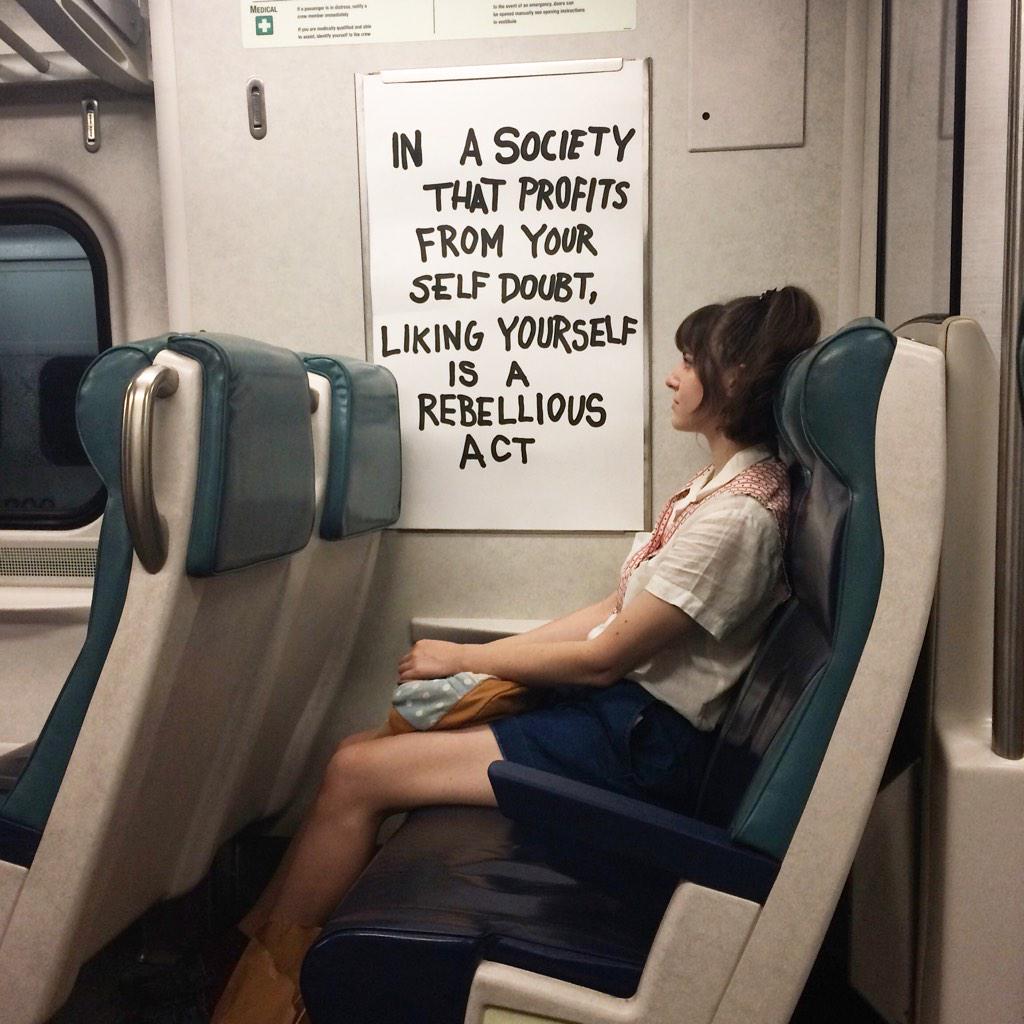 Woman sitting on a train next to a sign that says: In a society that profits from your self doubt, liking yourself is a rebellious act.